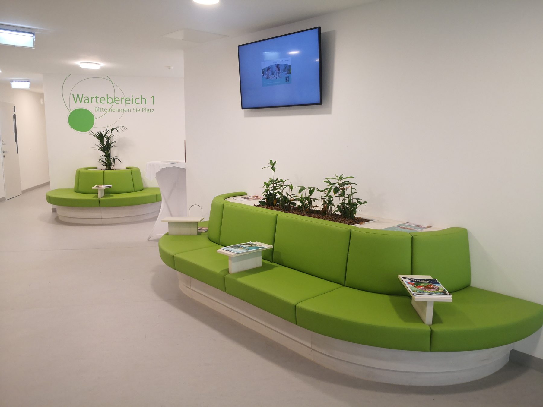 Wartebereich der Ordination mit grünen Sitzbänken, Pflanzen und Monitor